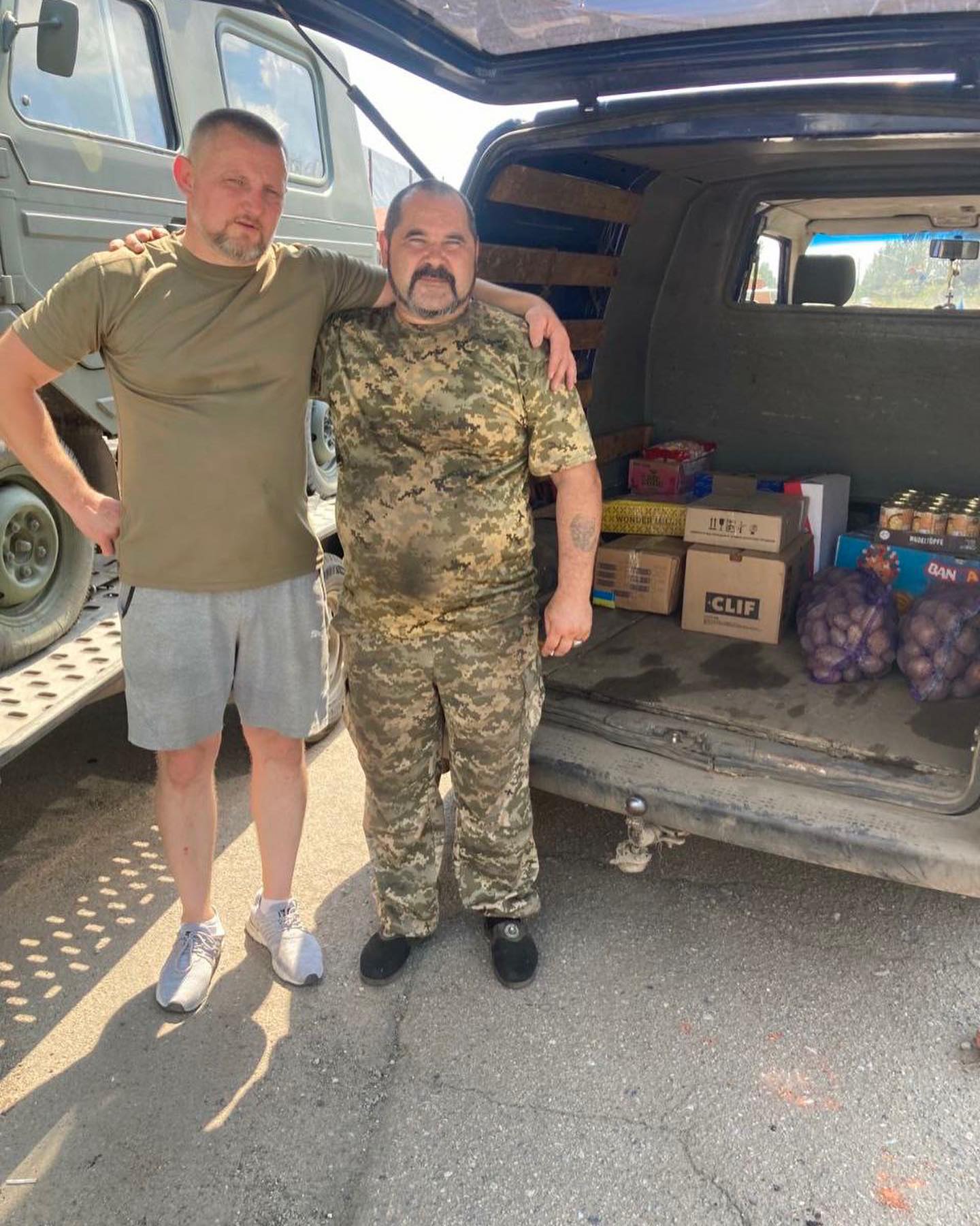 two men standing next to a van full of food.
