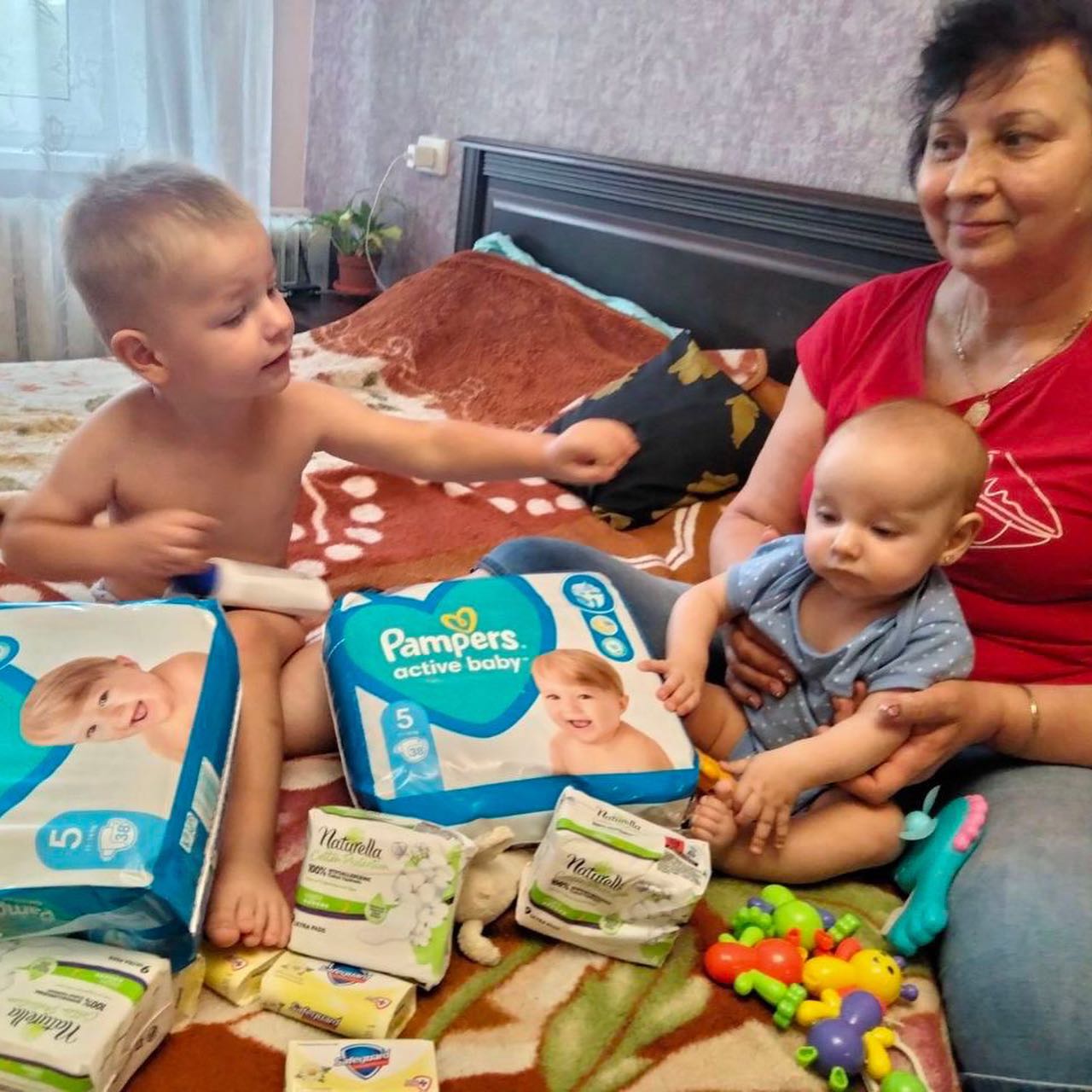 A woman and two children sitting on a bed with packs of pampers diapers.