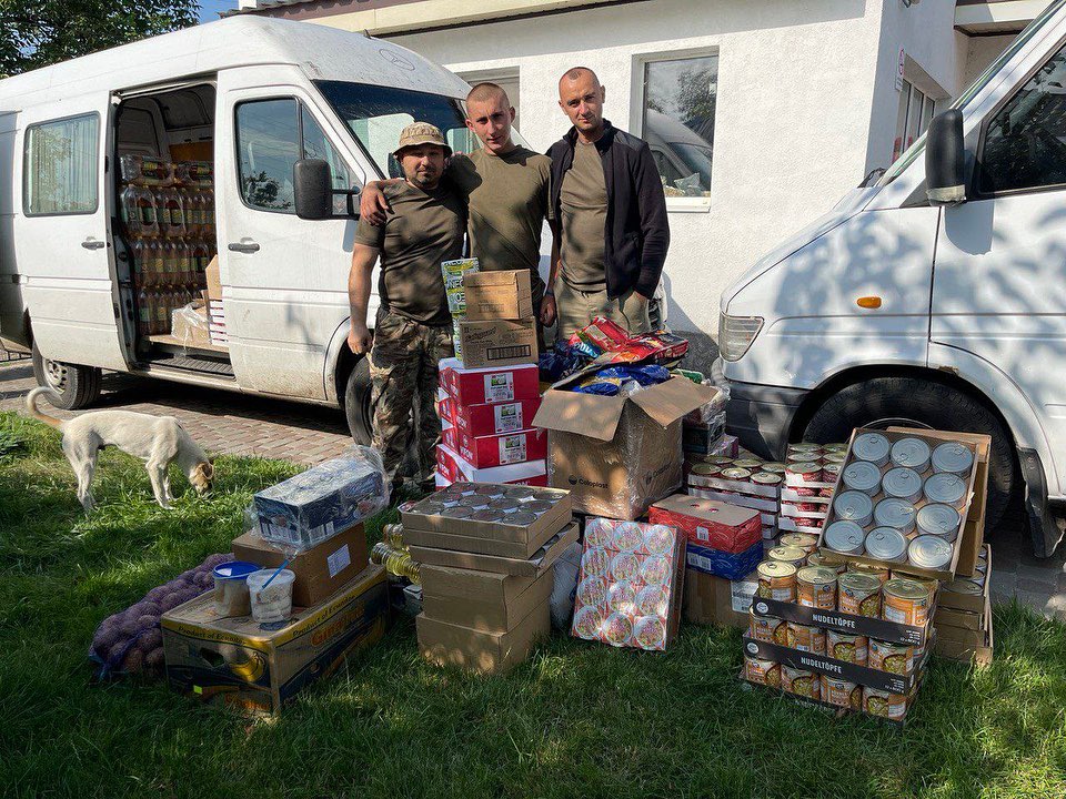 Three men standing in front of a van with boxes of food.