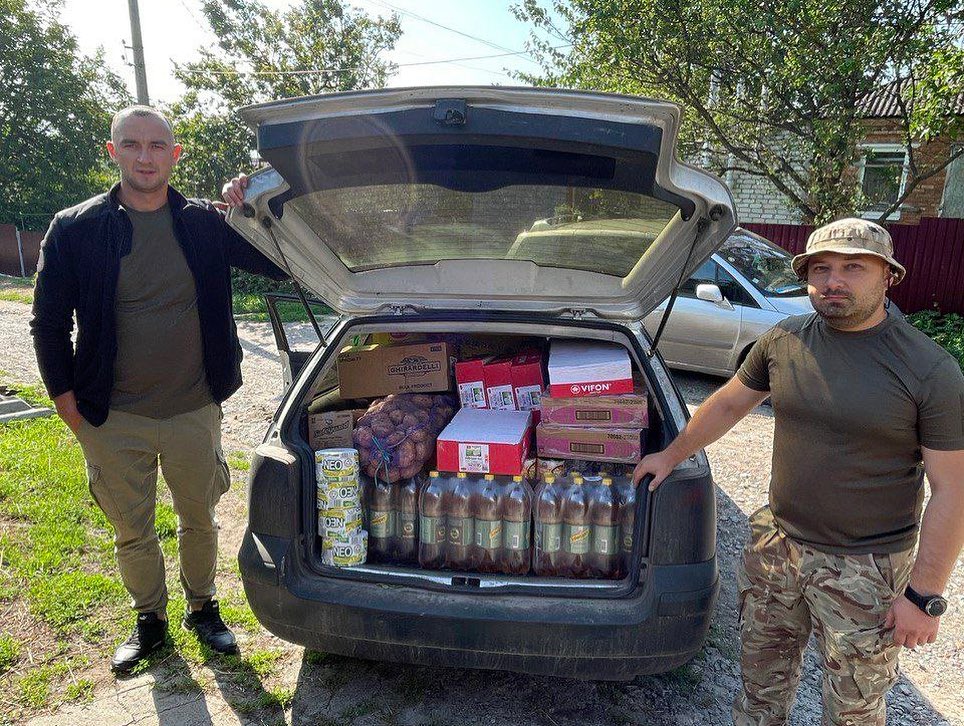 Two men standing next to a car with a trunk full of food.