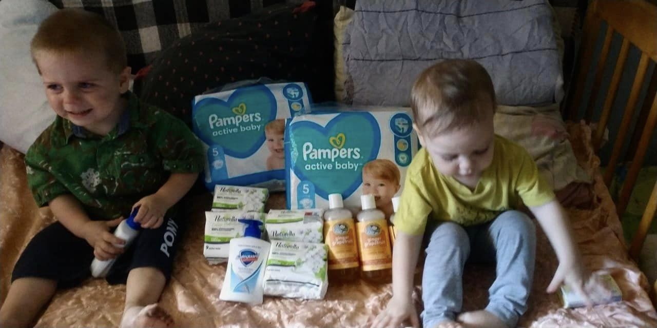Two boys sitting on a bed with a bunch of baby products.