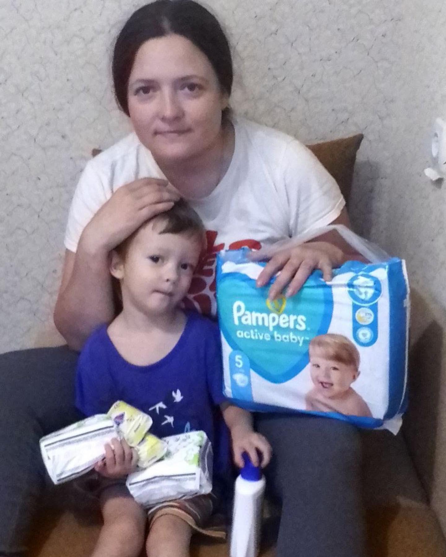 A woman and child sitting on a couch with a pack of pampers diapers.