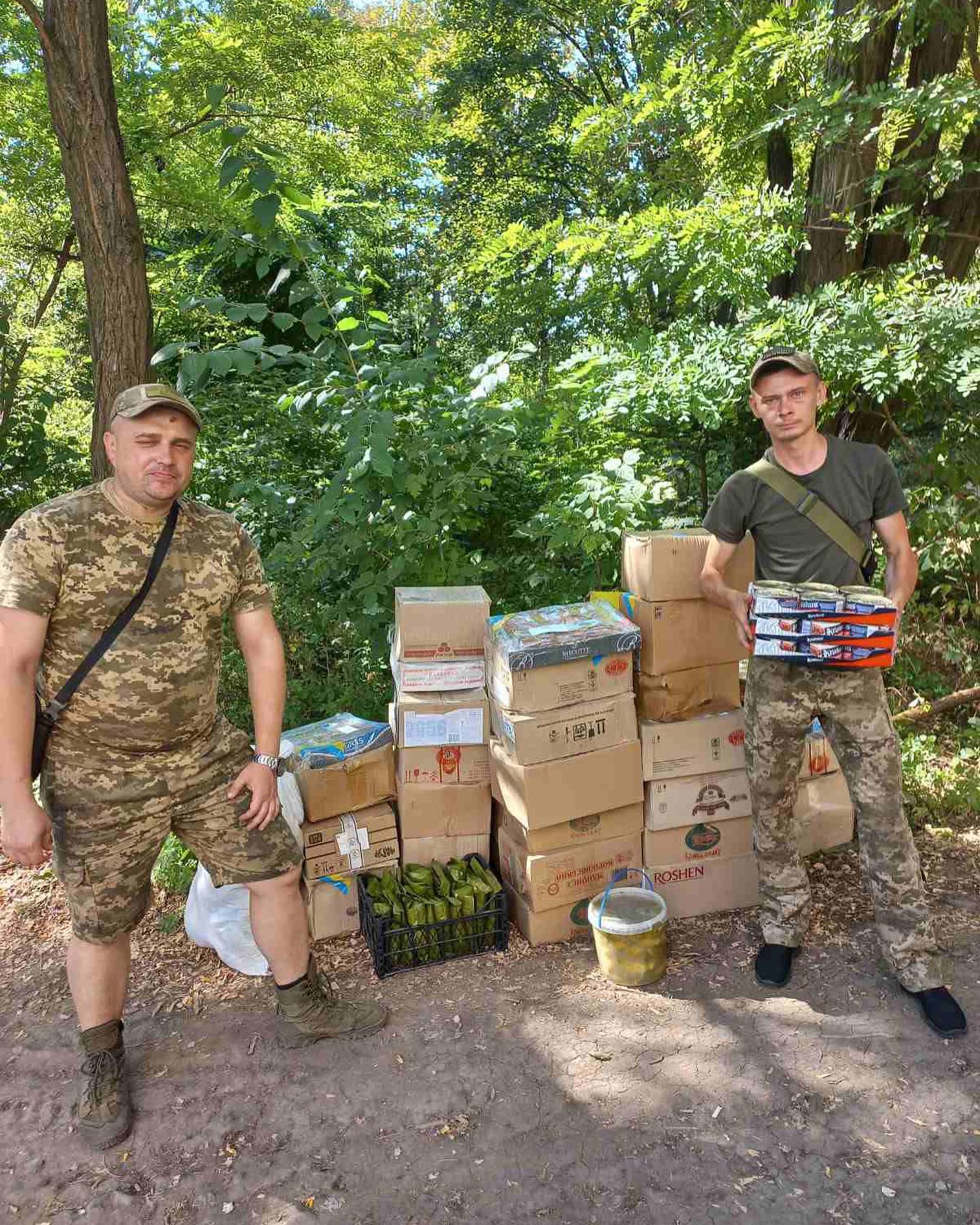 Two men in camouflage standing next to boxes of food.