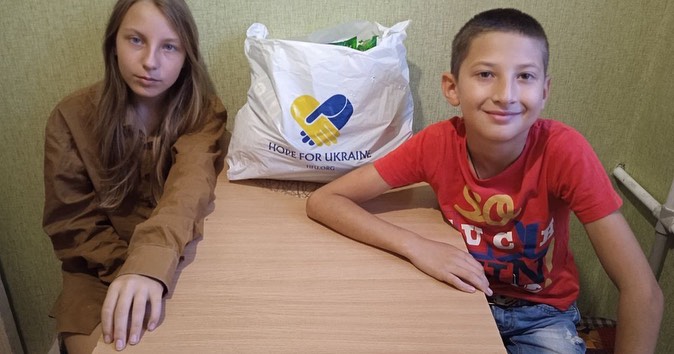 Two children sitting at a table with a bag in front of them.