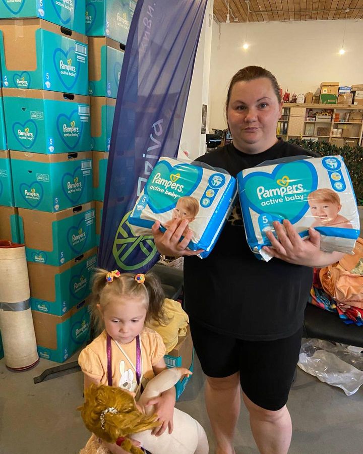 Two women and a little girl standing in front of boxes of diapers.