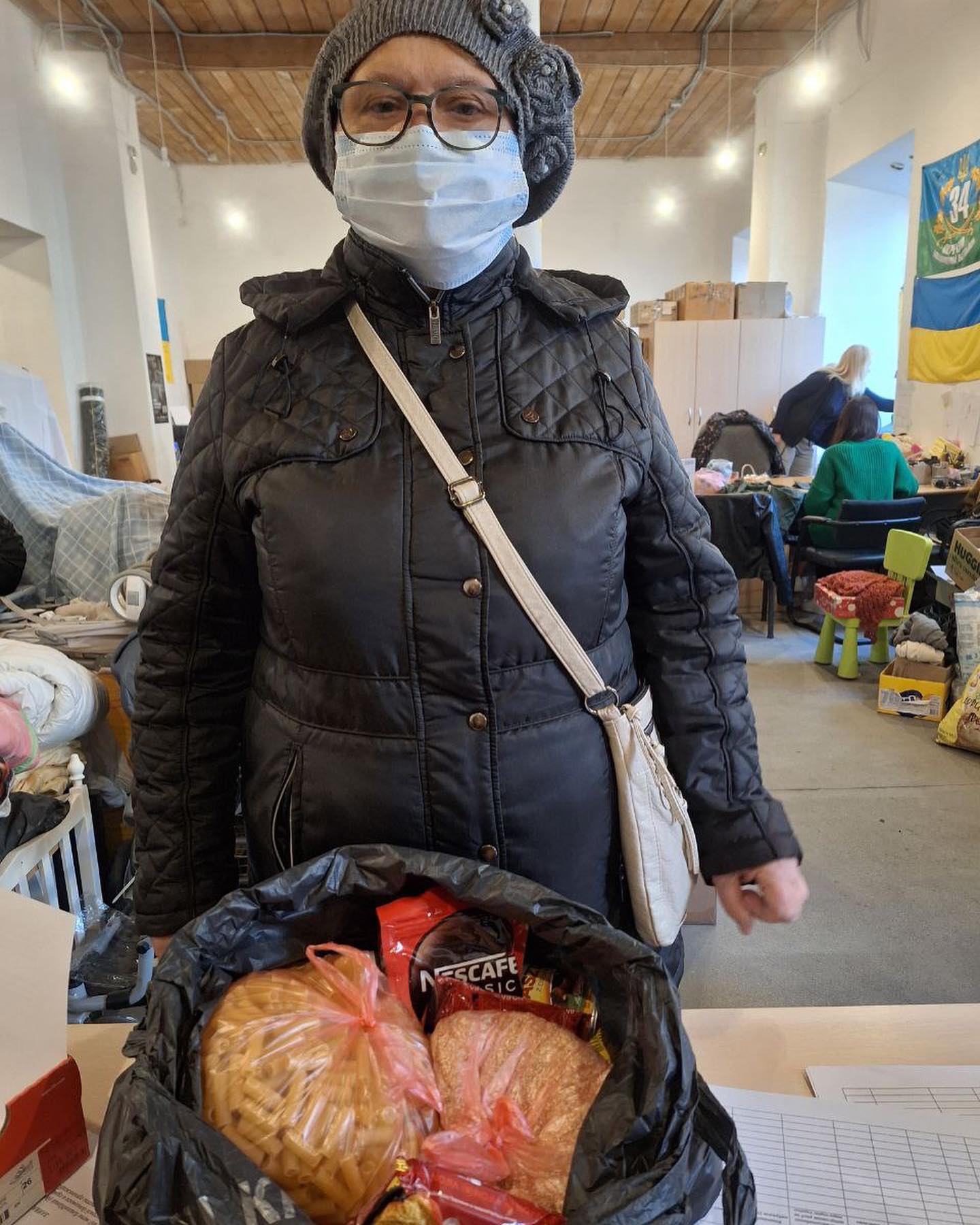 A woman wearing a face mask stands next to a bag of food.