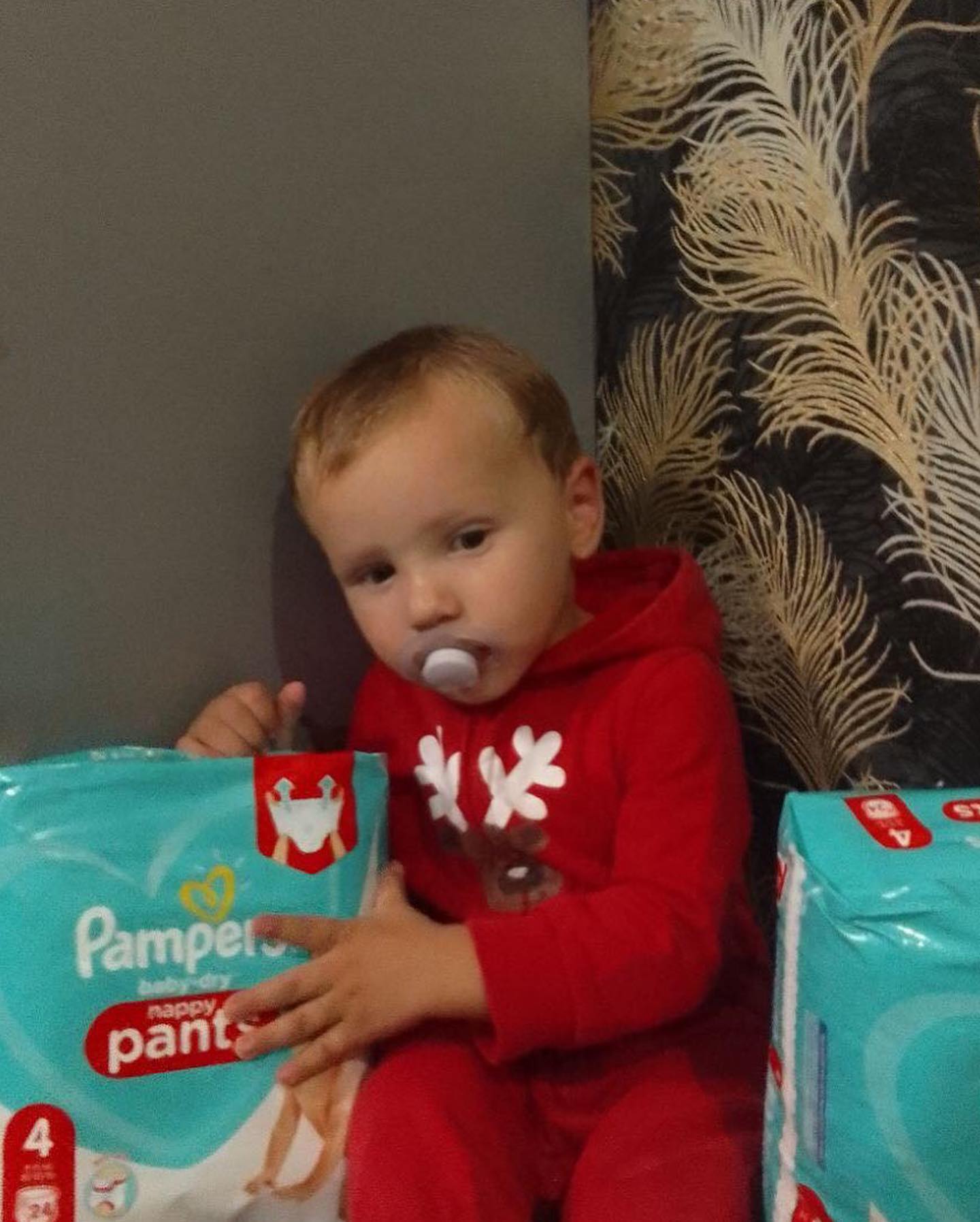A young boy in a red pajama sitting next to a pack of pampers pajamas.
