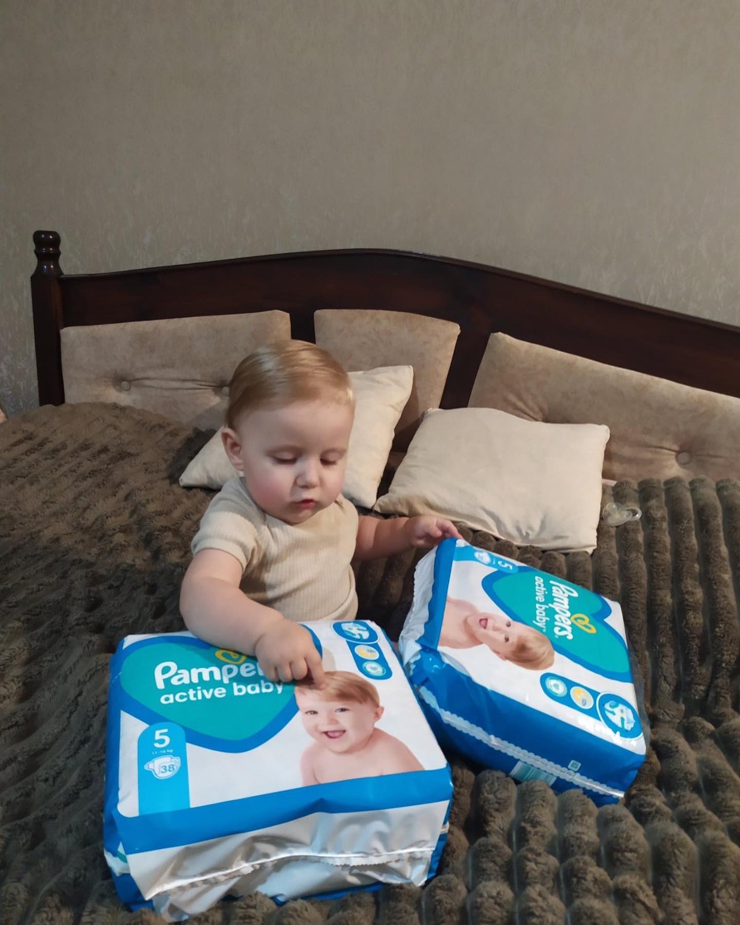 A baby sitting on a bed with two boxes of pampers diapers.