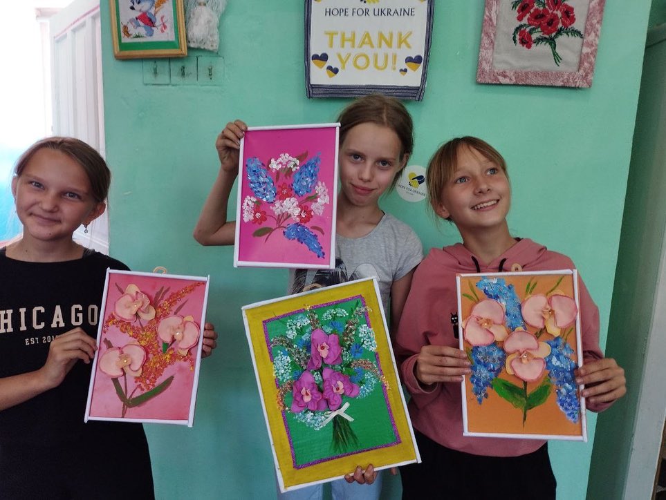 Three girls holding up flower paintings in a room.