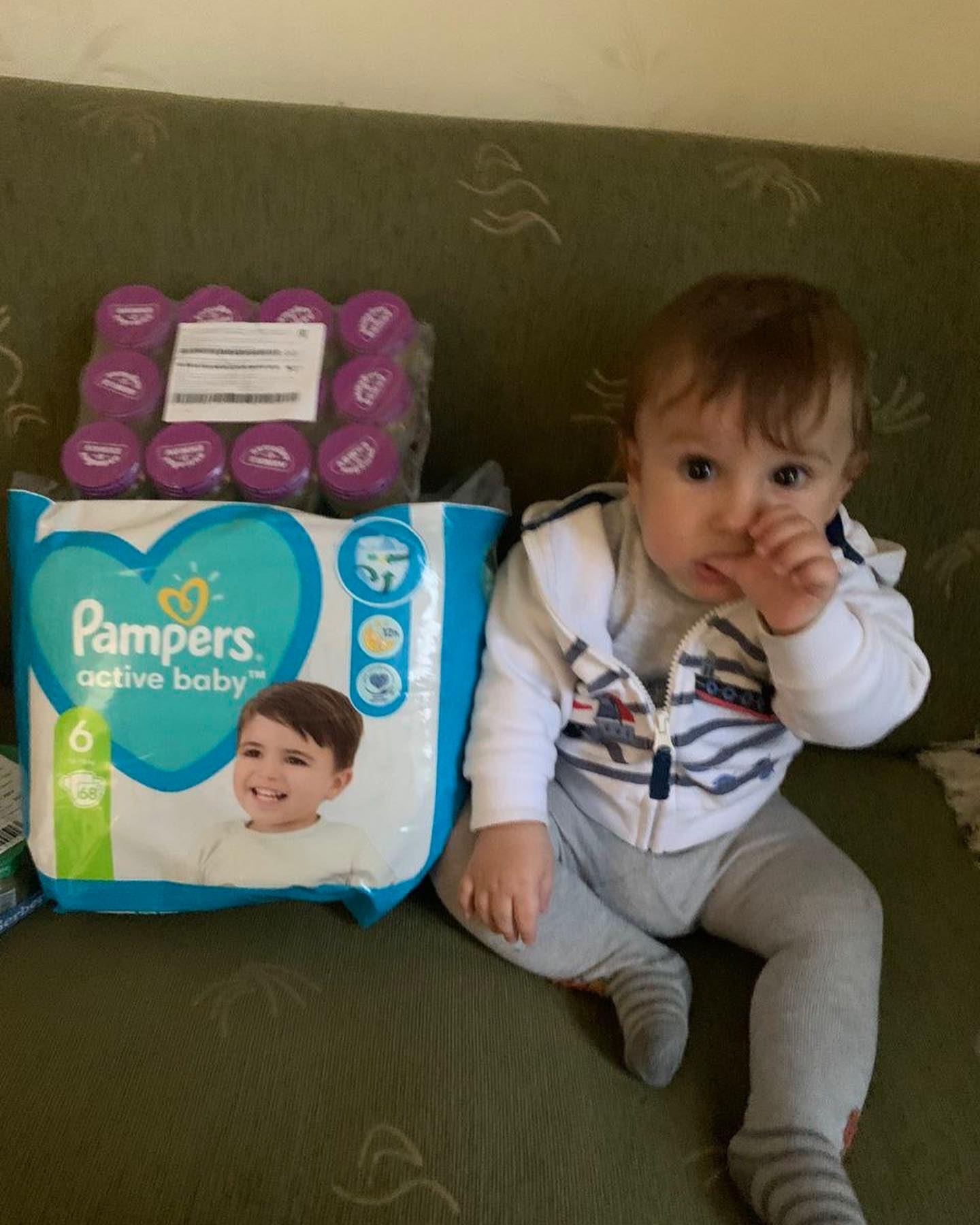 A baby sits on a couch next to a box of pampers diapers.