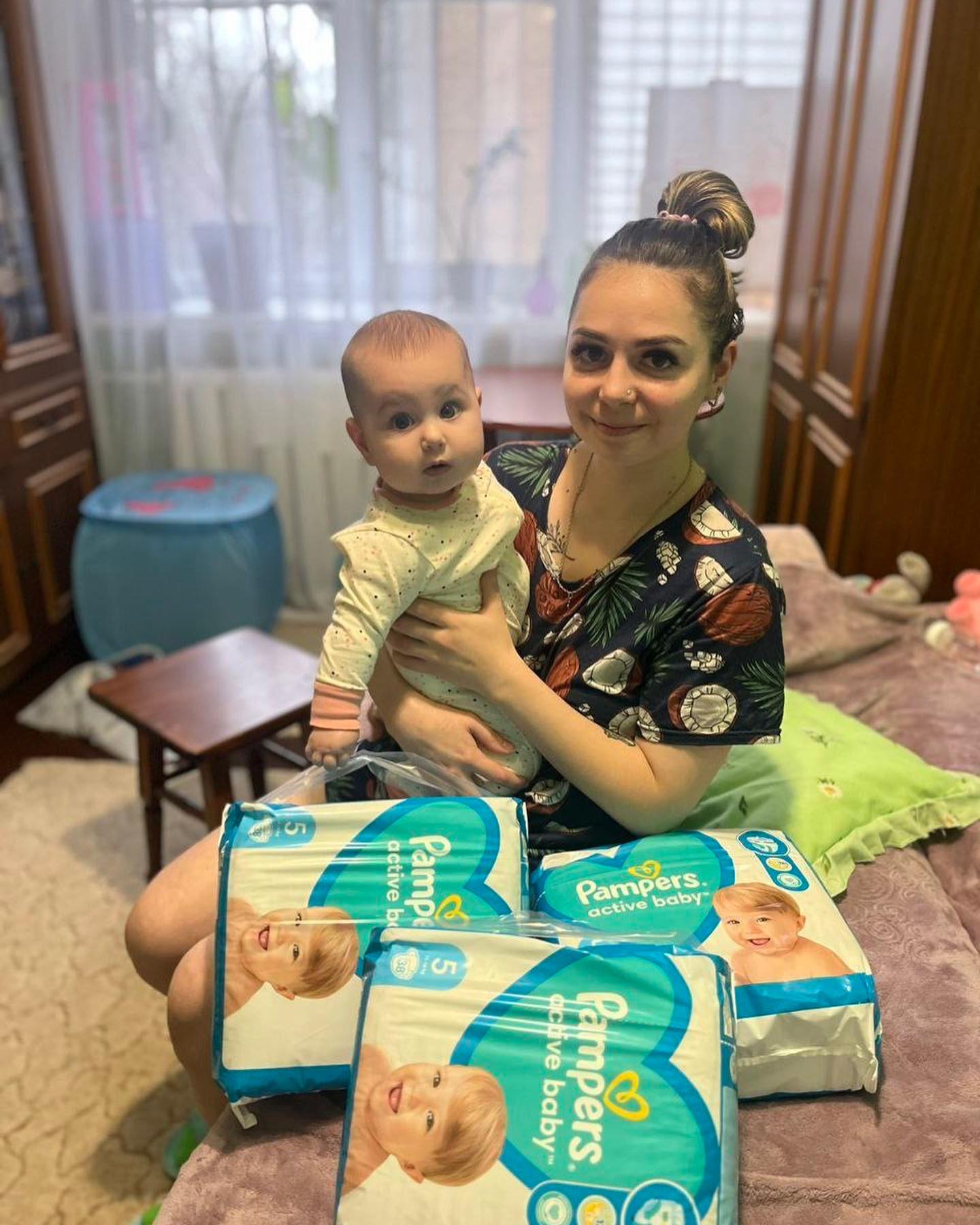 A woman with a baby sitting on a bed with packs of pampers diapers.