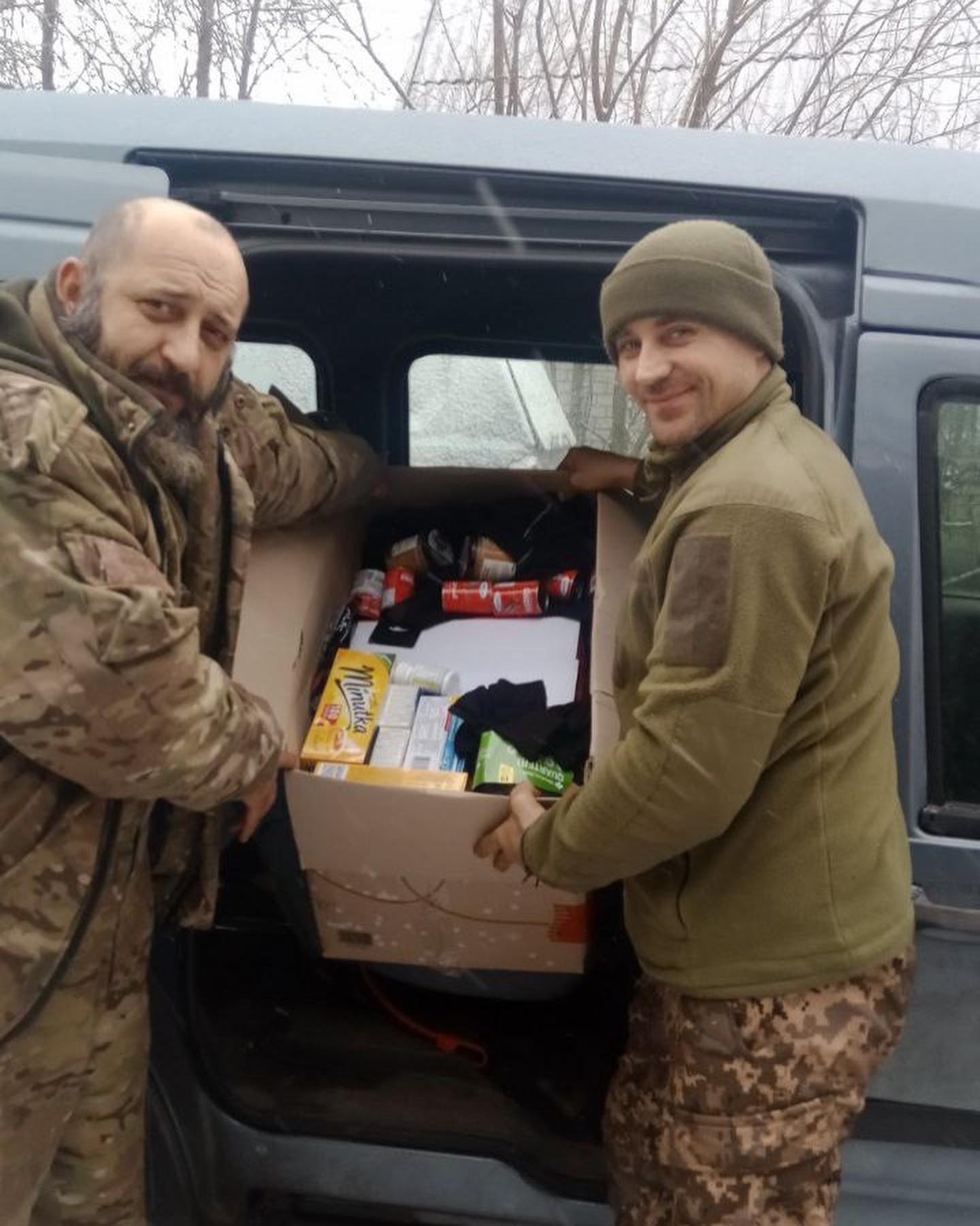 Two men in camouflage standing next to a van full of food.
