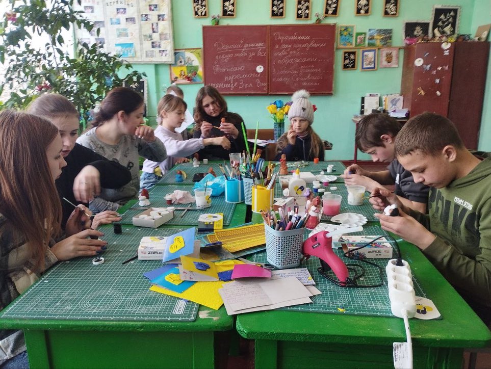 A group of students engaged in a craft activity at a classroom table, surrounded by art supplies and colorful materials.