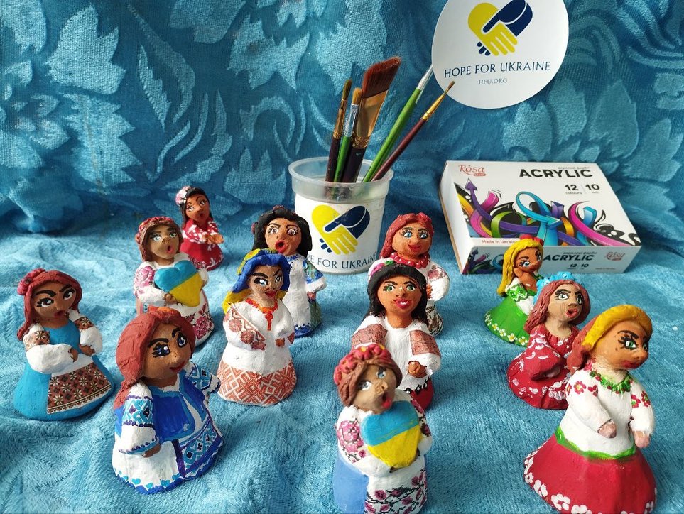 A collection of colorful, hand-painted figurines displayed in front of a "hope for ukraine" sticker, alongside paintbrushes and acrylic paints.