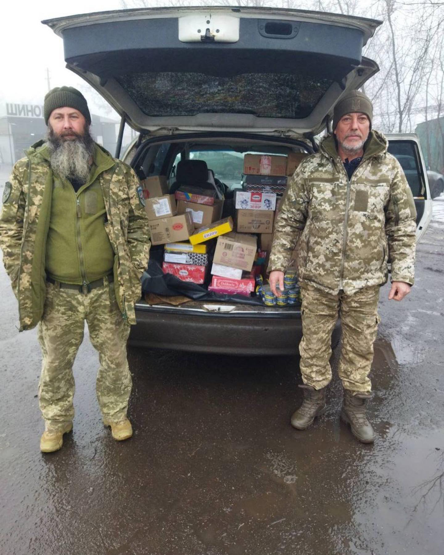 Two men in camouflage stand by an open car trunk full of supply boxes, on a foggy day.