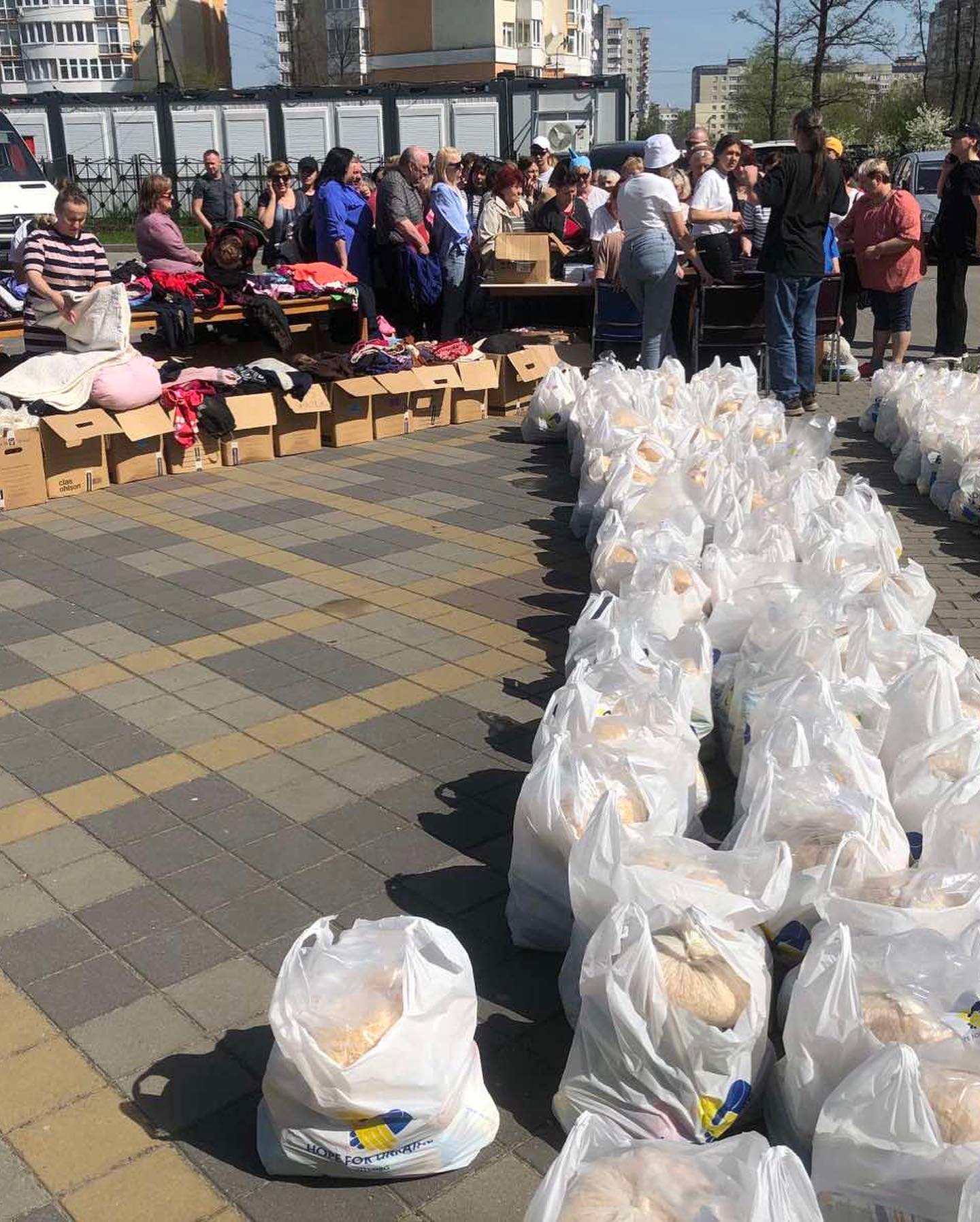 A group of people gathering around tables with clothes and boxes, with rows of plastic bags filled with goods in the foreground.
