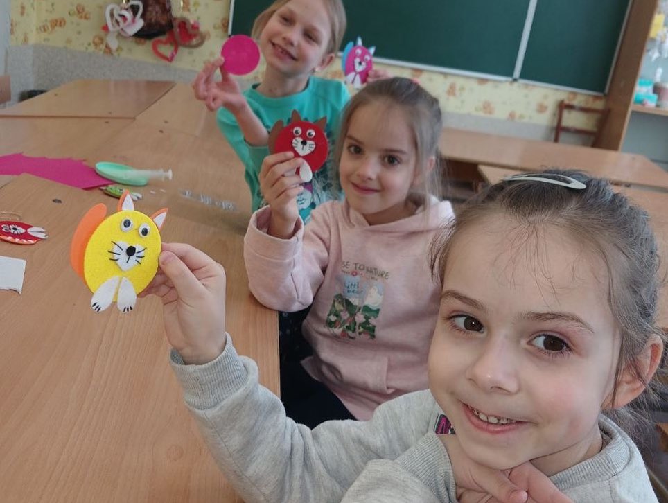 Three children smiling at the camera, displaying colorful paper crafts they made in a classroom.