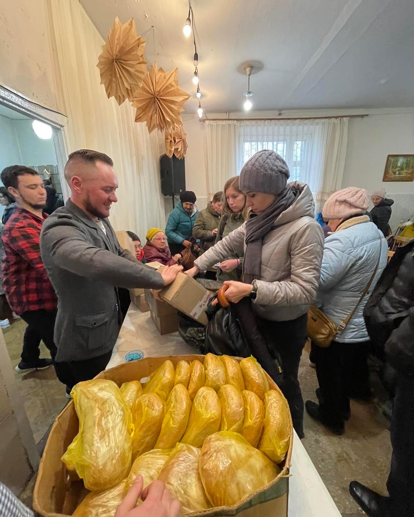 A man hands out bread to a woman in a crowded indoor setting with people waiting in line for humanitarian aid.