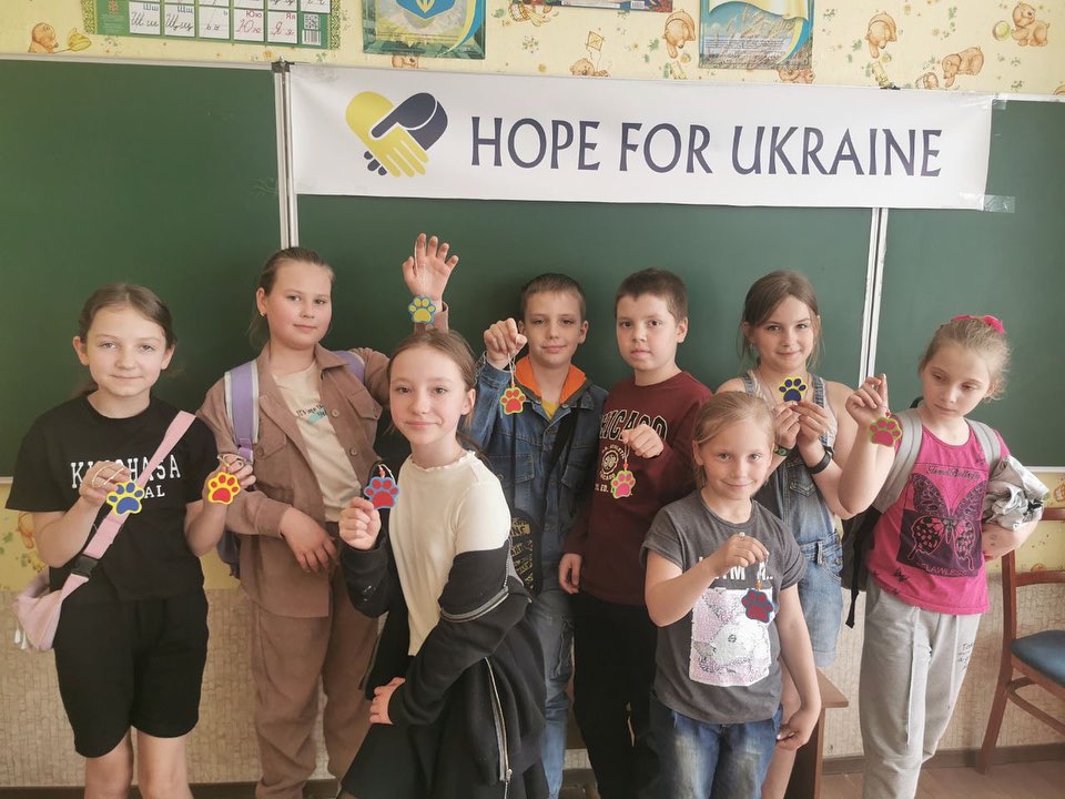 Eight children standing in a classroom, smiling and holding colorful crafts, with a banner reading "Support Ukraine" in the background.