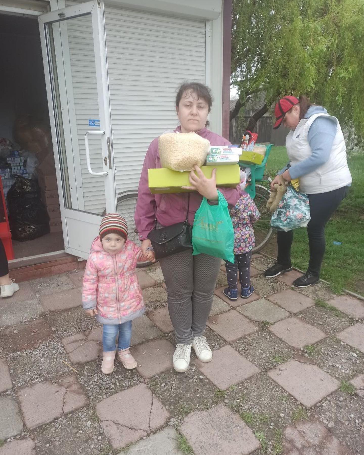 A woman holding a large loaf of bread and a box of humanitarian aid stands outside a garage with a young child by her side, as another person works in the background.