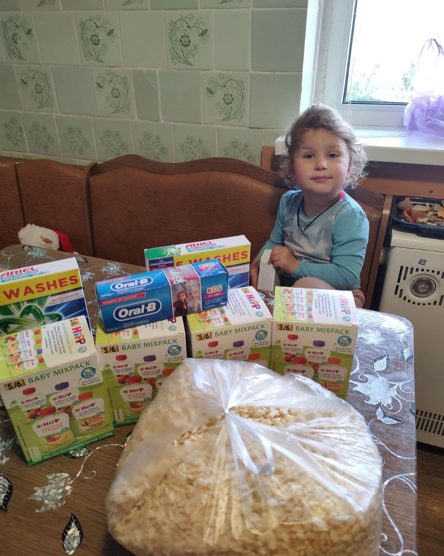 A young child sitting at a kitchen table surrounded by various household and personal care products, including toothpaste and baby wipes, collected for Ukraine relief efforts.