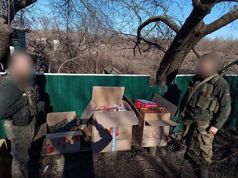 Two blurred-faced soldiers standing beside cardboard boxes filled with humanitarian aid, outdoors near a green fence and leafless trees.