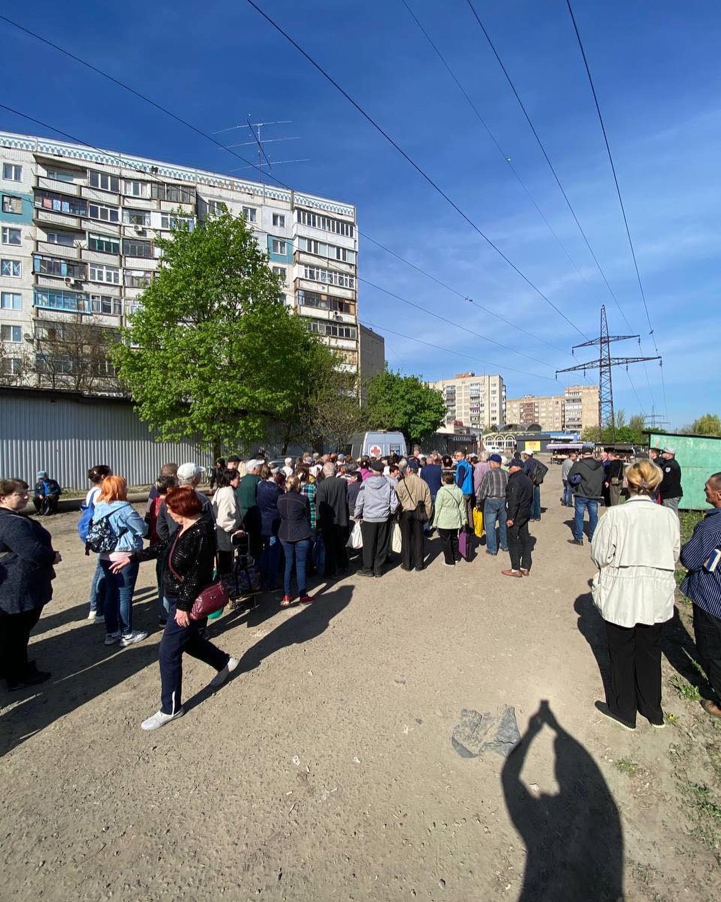 Crowd of people waiting in a line on a dirt path with residential buildings in the background under a clear sky, hoping for Ukraine aid support.