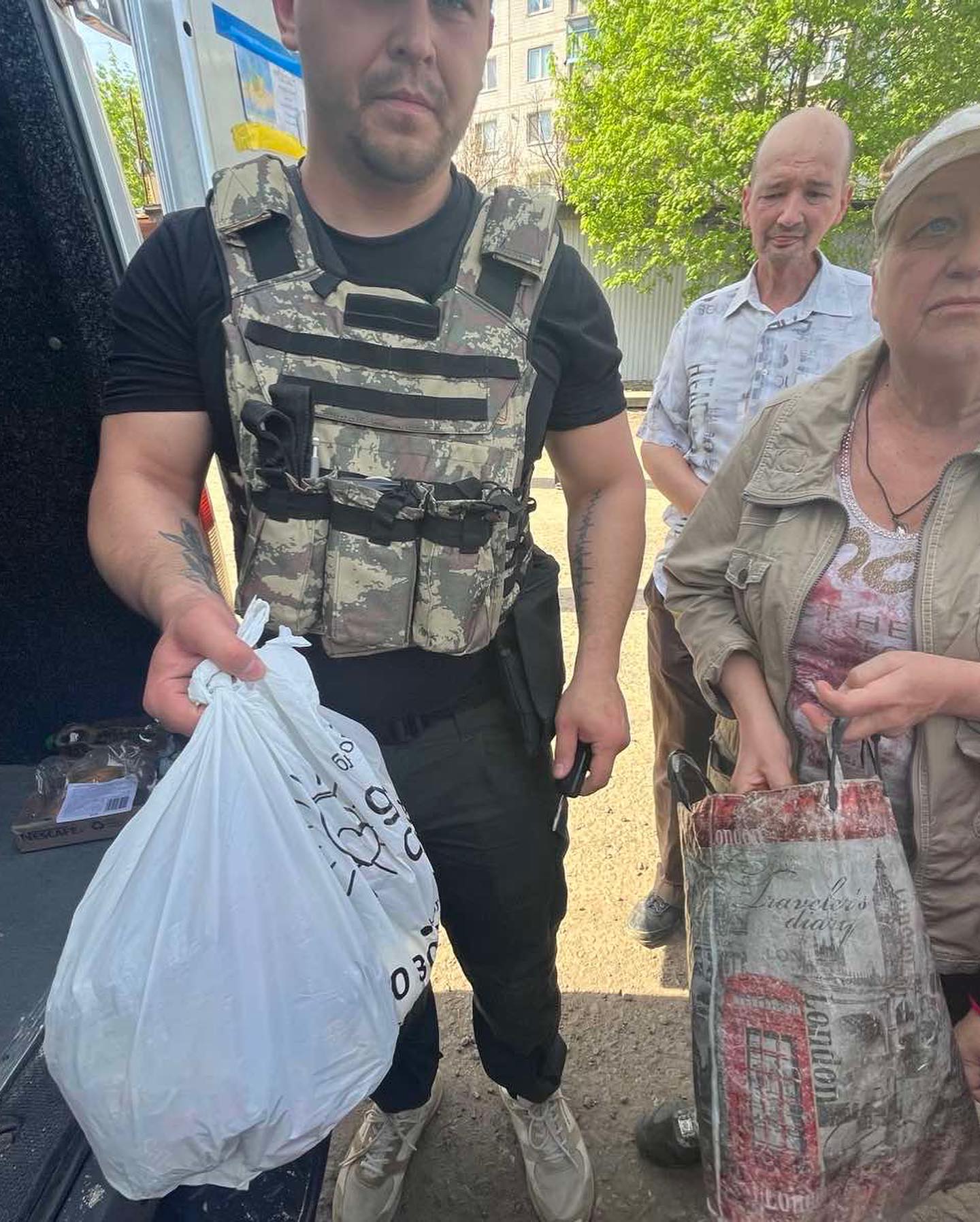 A volunteer in a tactical vest hands a plastic bag to an elderly woman near a van, with another man observing in the background.