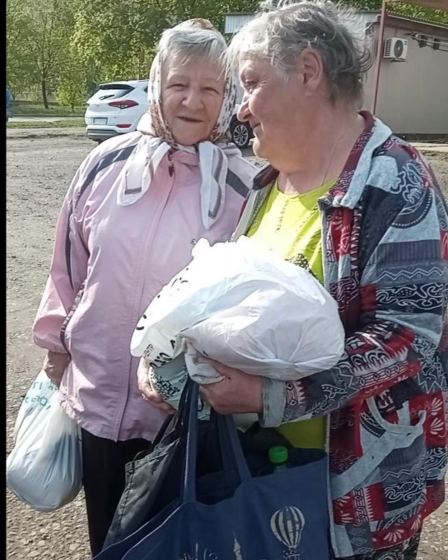 Two elderly women chatting outdoors, one holding plastic bags with supplies for Ukraine aid, surrounded by cars and greenery.