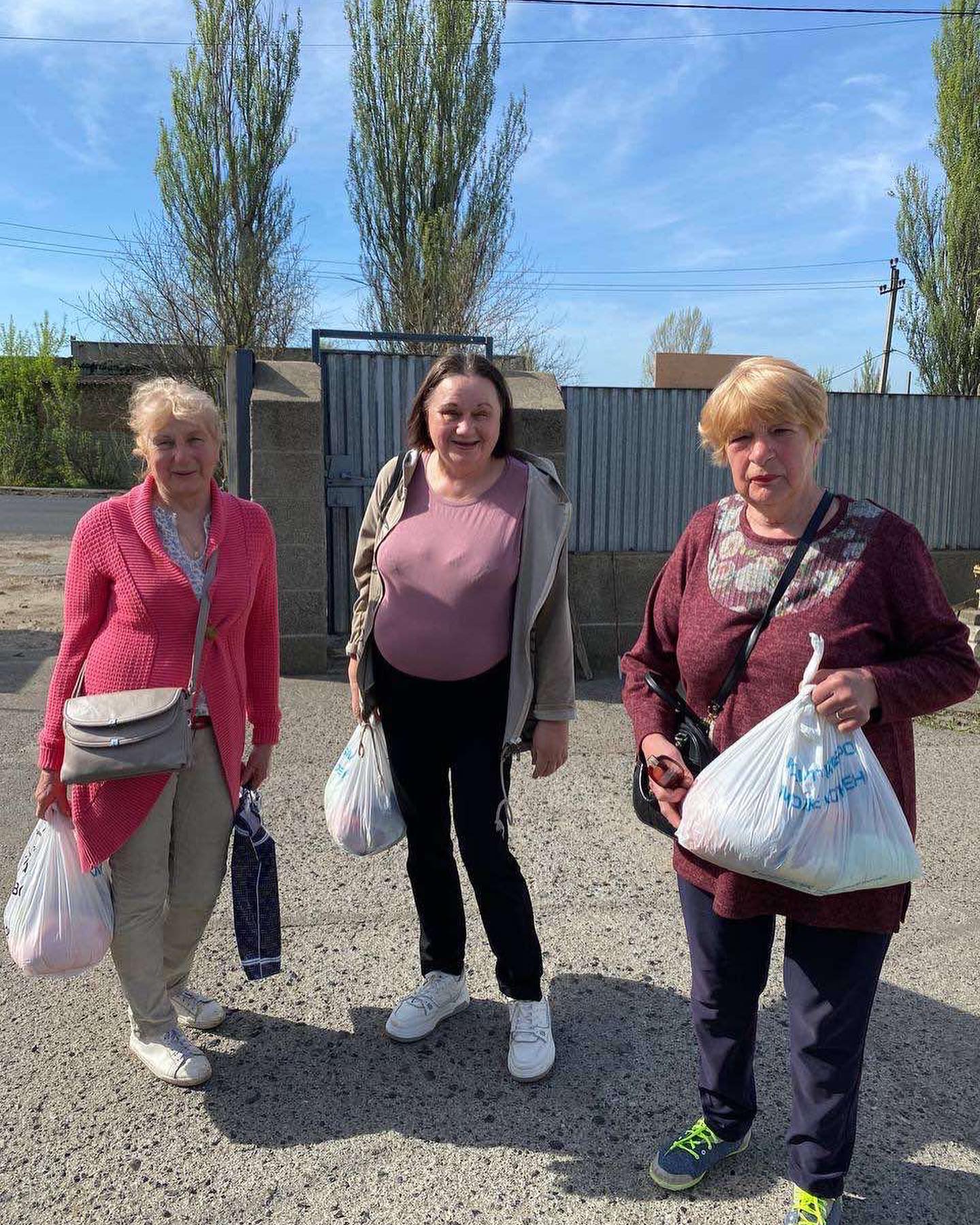 Three women standing outside, smiling, each holding shopping bags. Two elderly, one middle-aged, in casual attire on a sunny day. They volunteer to help Ukraine.