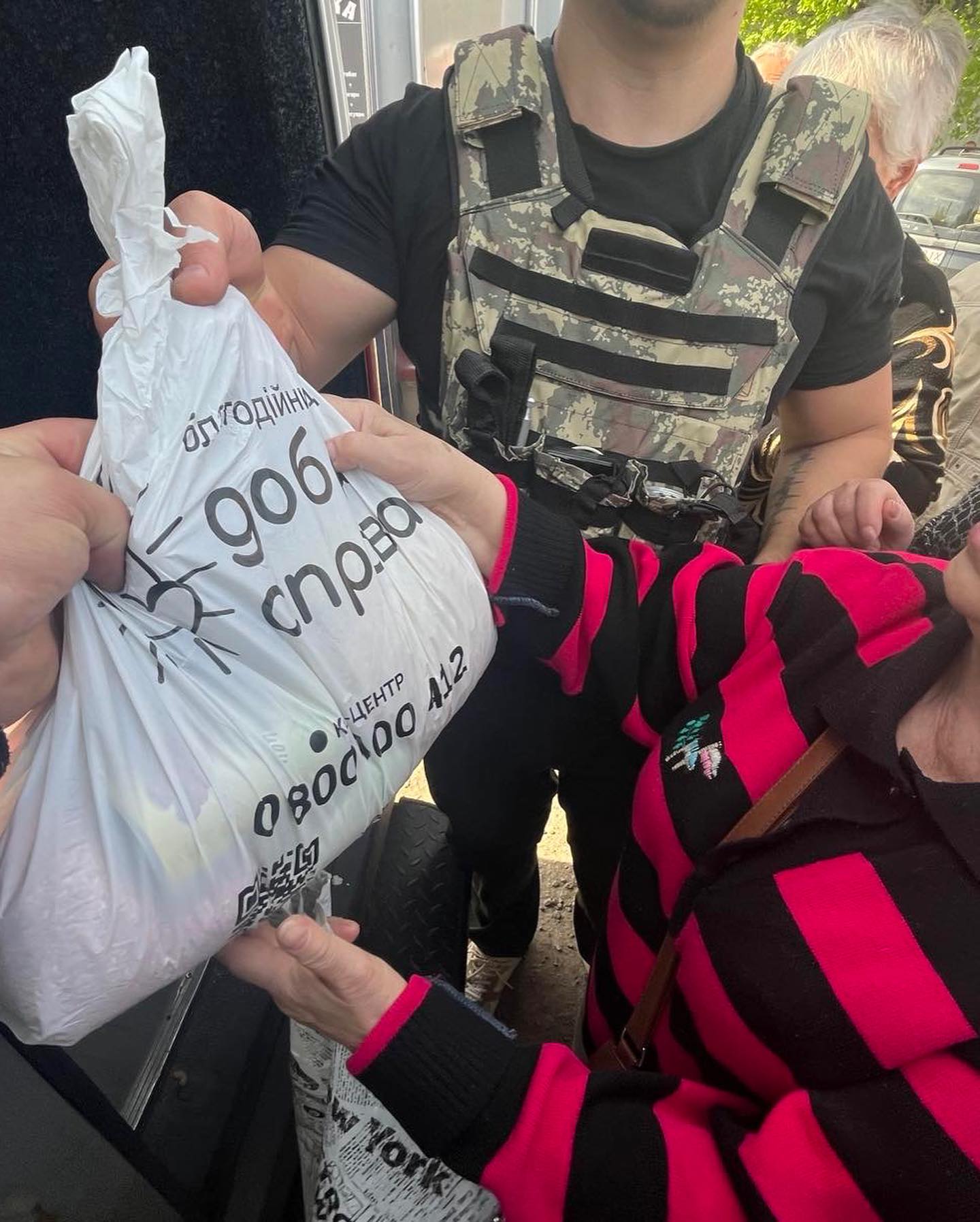 A person in a bulletproof vest hands a plastic bag marked in Cyrillic, containing humanitarian aid, to another person in a pink and black striped jacket.