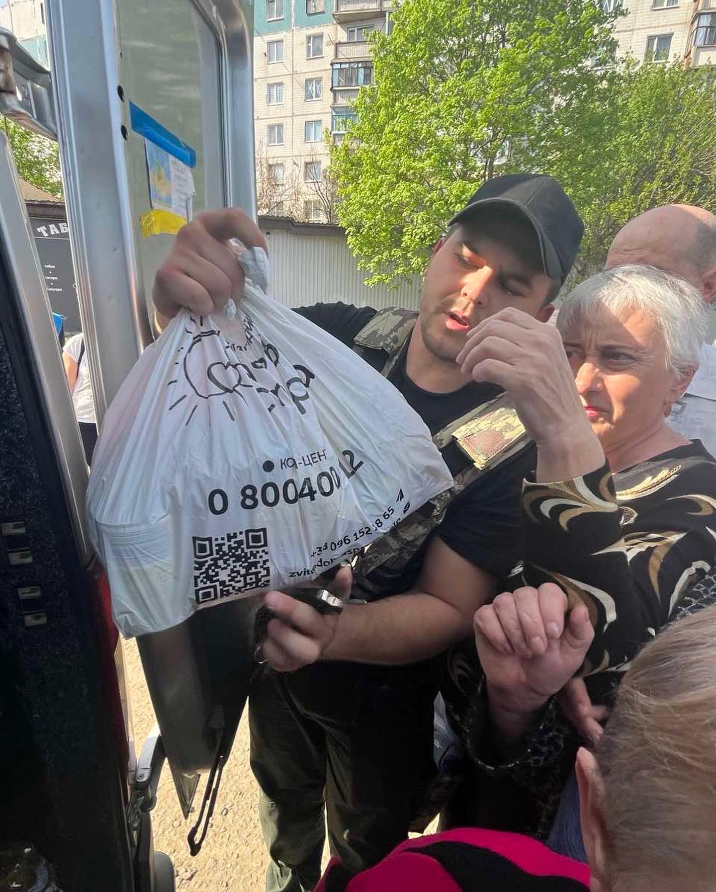 A young man hands a plastic bag labeled "Hope for Ukraine aid" and a QR code to an elderly woman by a bus, as other passengers look on.