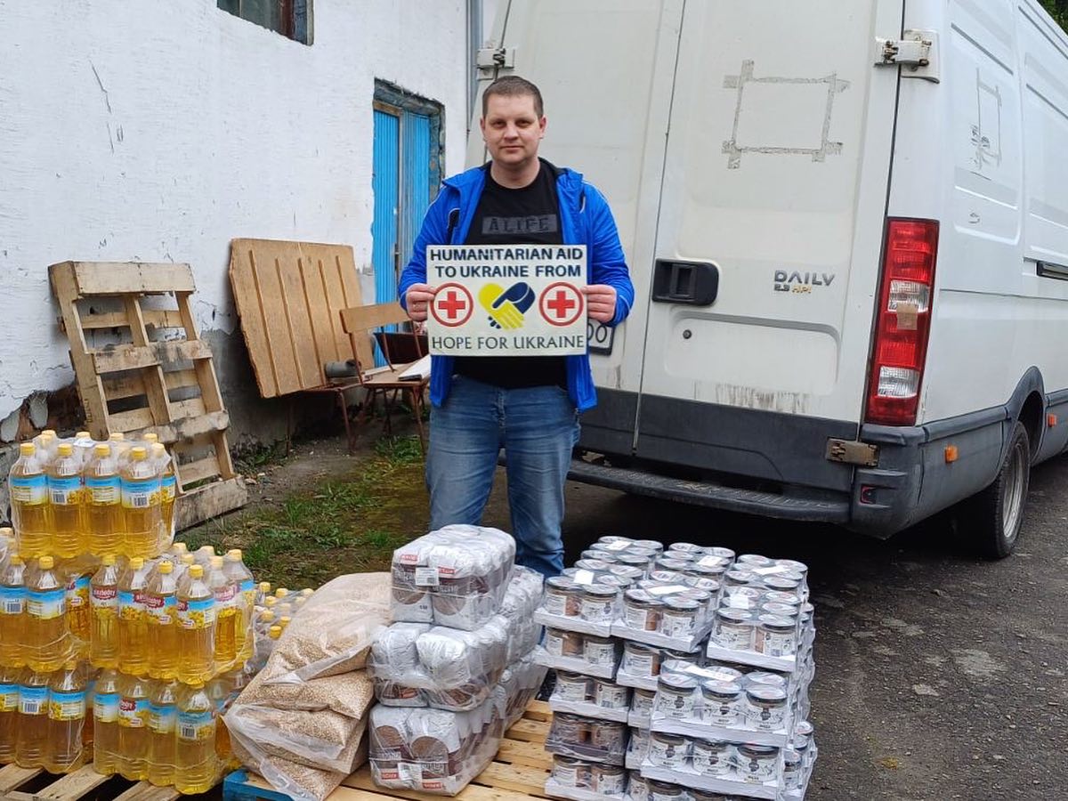 A man stands in front of a van and pallets loaded with bottled water and food supplies, holding a sign reading “humanitarian aid to Ukraine from [Hope for Ukraine Support logo].”