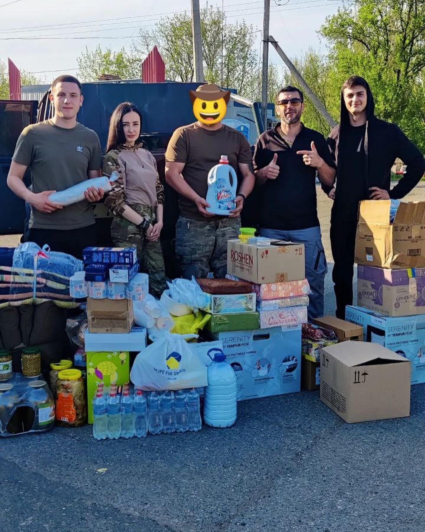Group of volunteers with supplies such as food, water, and household items, smiling and posing for a photo outdoors in support of Ukraine relief efforts. One person's face is covered with a smiley face