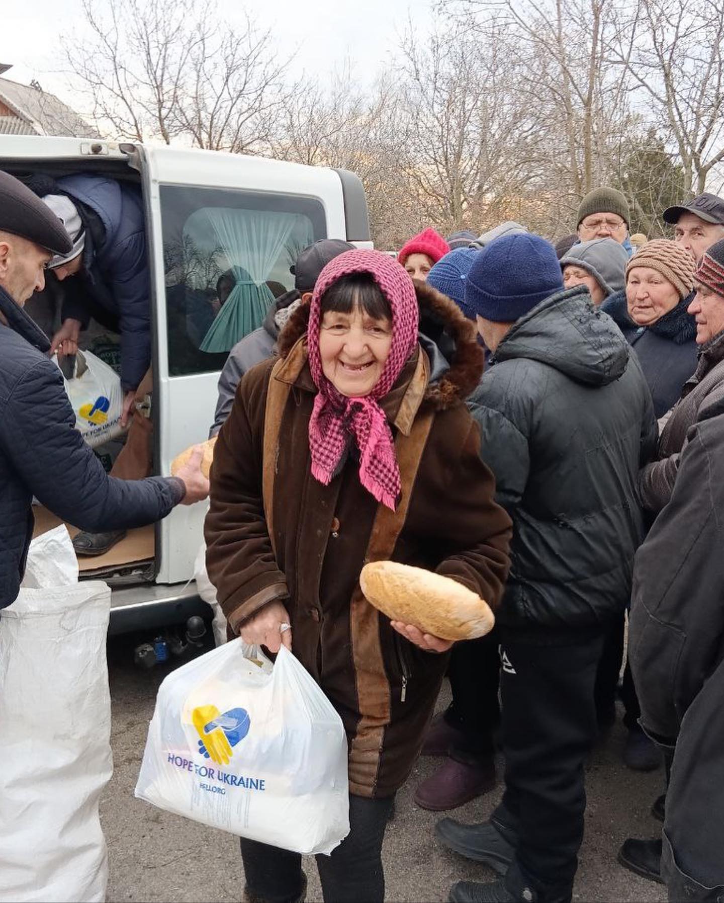 An elderly woman in a brown coat and pink headscarf smiles while receiving bread and supplies from a volunteer. a van marked "hope for ukraine" is visible in the background.
