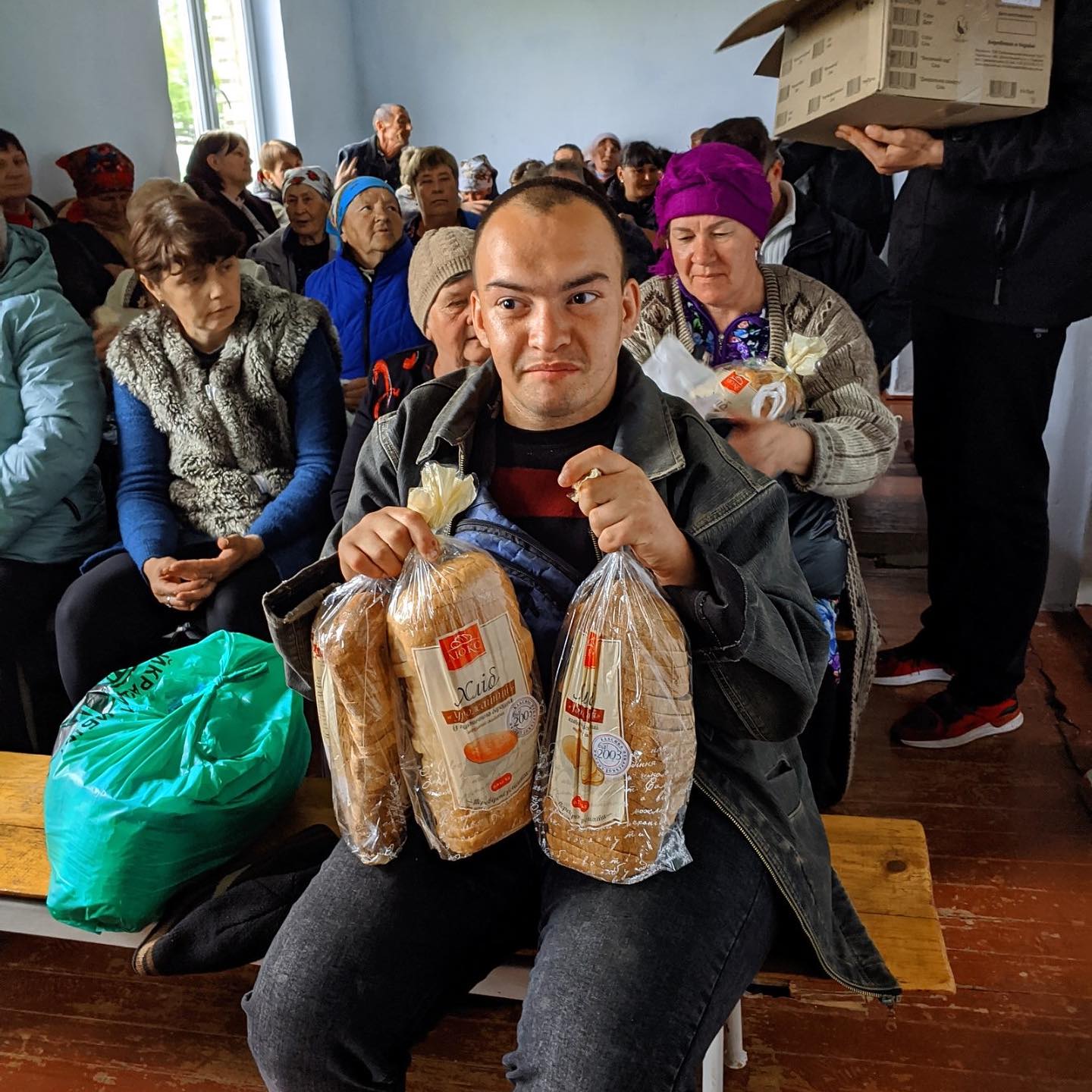 A young man sits on a bench holding two loaves of bread, surrounded by a group of seated displaced Ukrainians in a room.