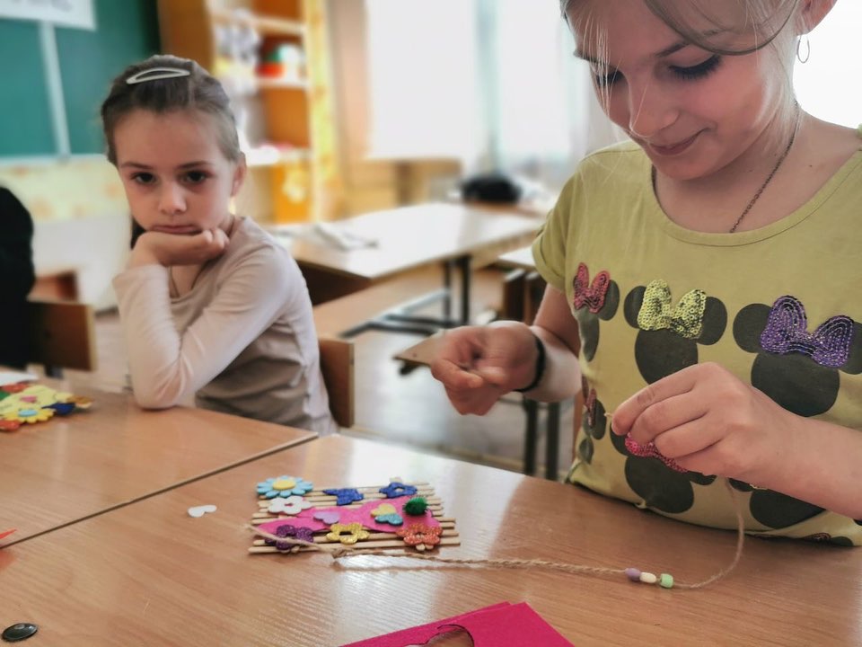 A woman instructs a young girl during a craft activity in the Donetsk region, threading beads, while the girl watches intently, arms crossed.