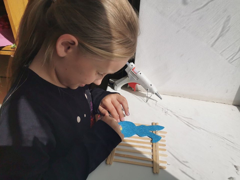 A young girl in the Donetsk region using a glue gun to apply glue to a blue hand-shaped cutout on a crafting table.