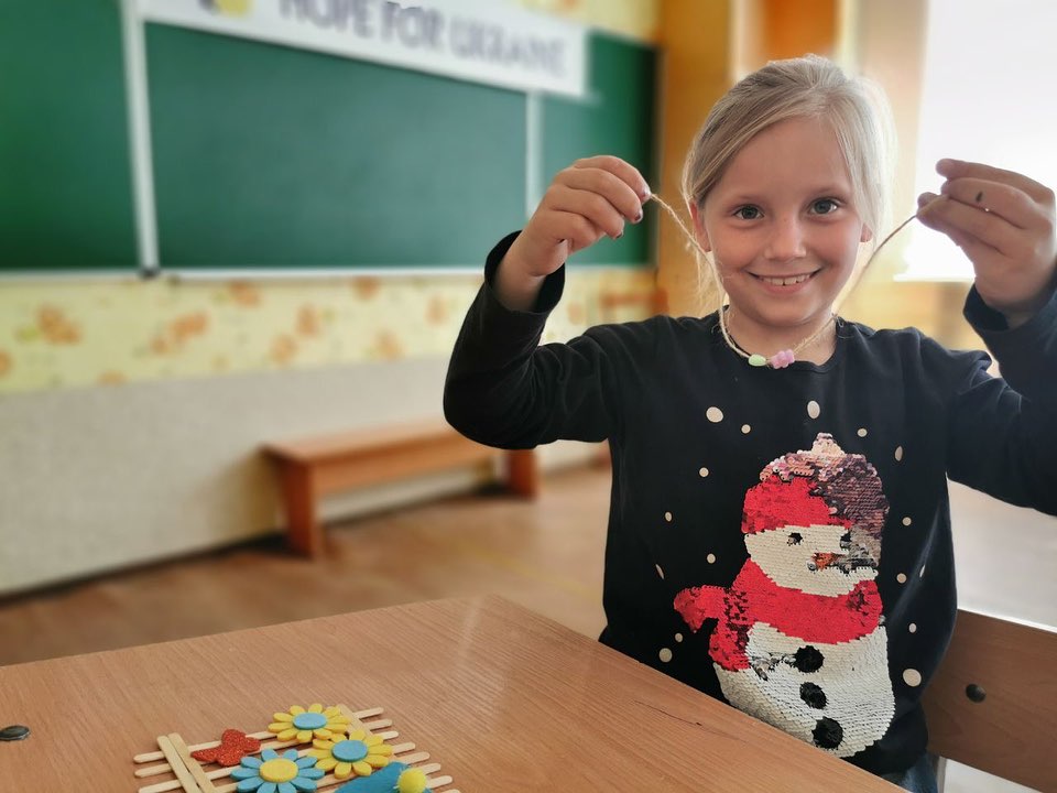A young girl with a blonde ponytail, wearing a snowman sweater, smiles while holding a craft project in a classroom in Donetsk region.