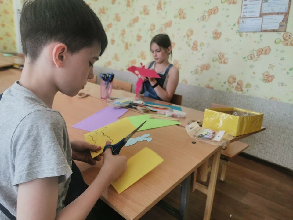 A boy cutting paper with scissors at a table while a woman crafts with paper in the background in a room, decorated with teddy bears, in the Donetsk region.
