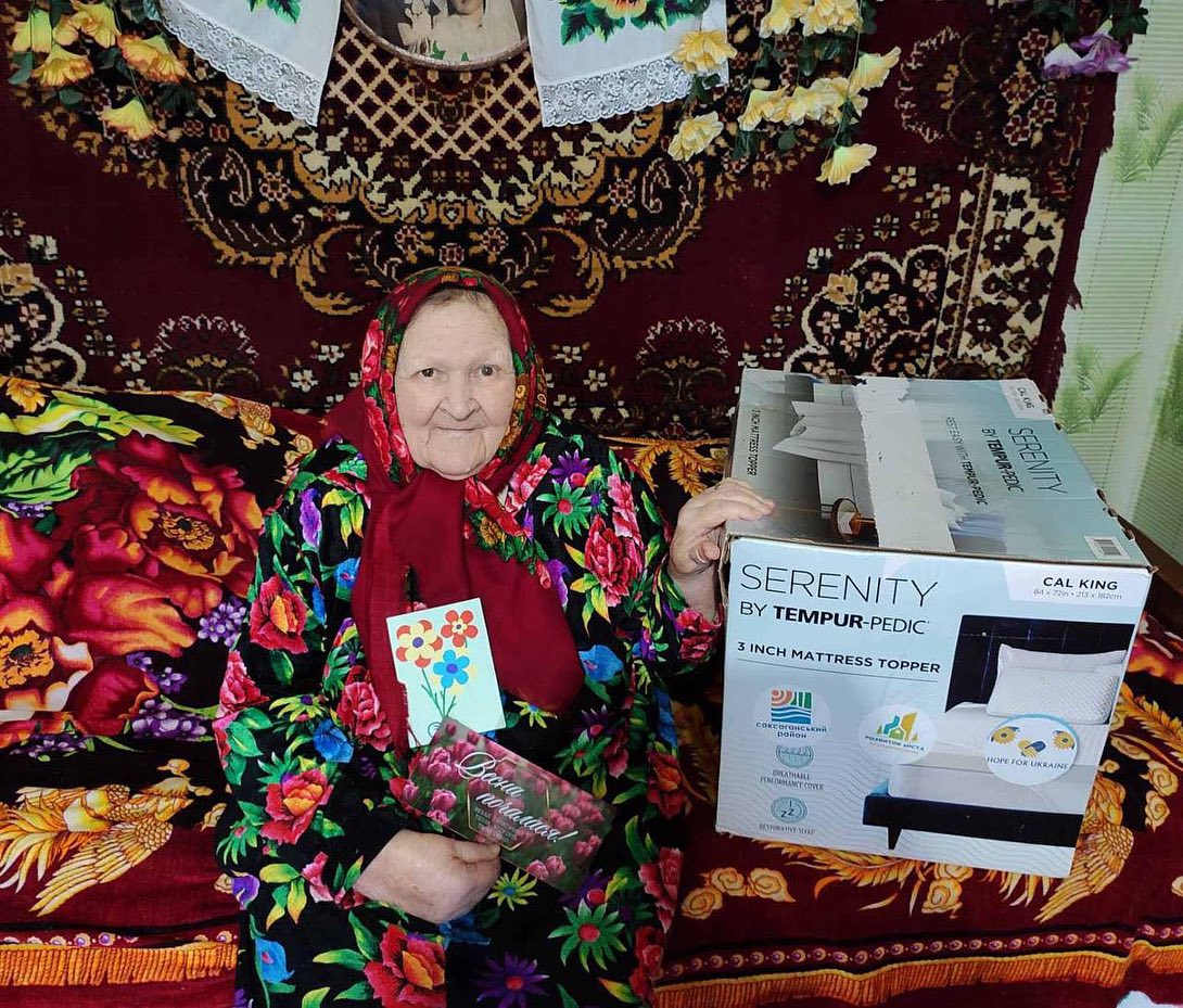 An elderly woman sitting on a couch, smiling and holding a flower-themed greeting card, with a new mattress topper in a box next to her.