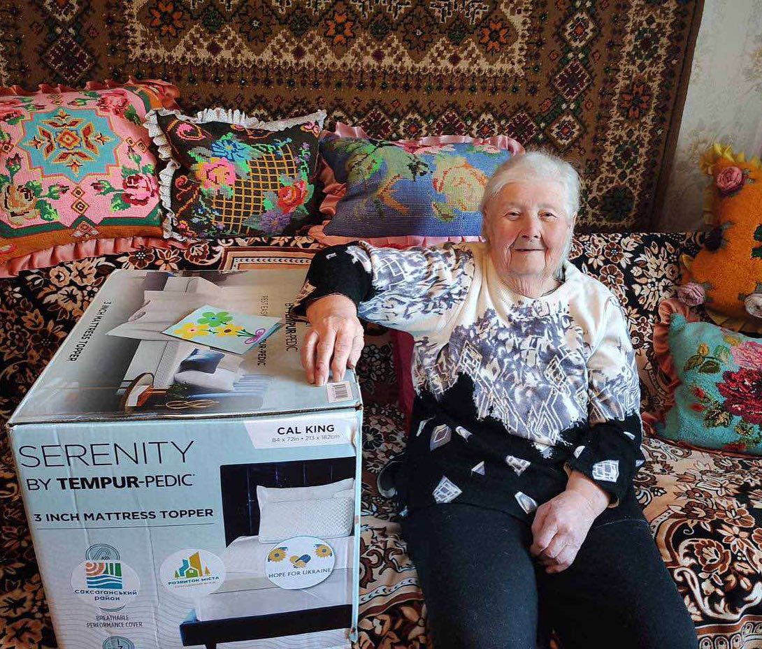 Elderly woman sitting beside a boxed mattress topper in a cozy, decorated room.