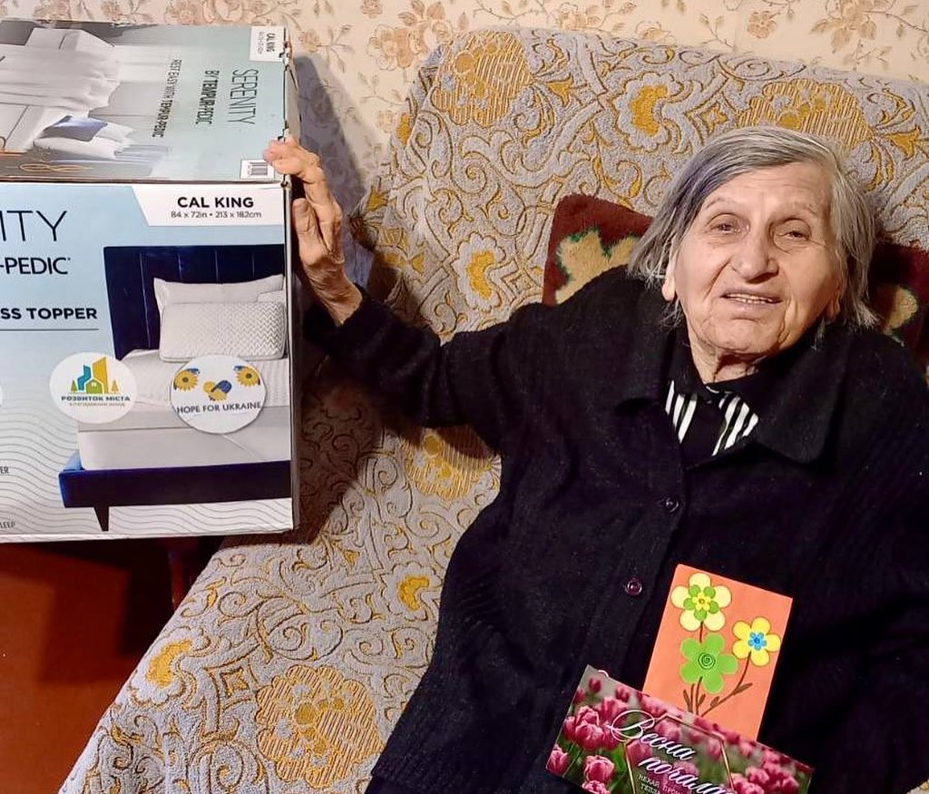 Elderly woman sitting on a couch, holding a mattress topper box, with a greeting card in her lap.