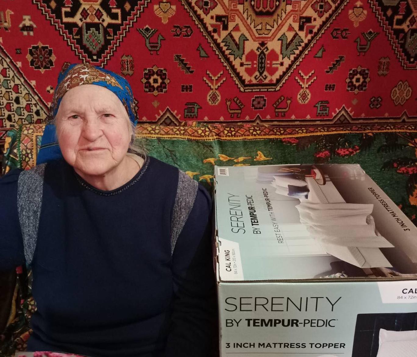 An elderly woman sitting in front of a colorful rug with a tempur-pedic mattress topper box beside her.