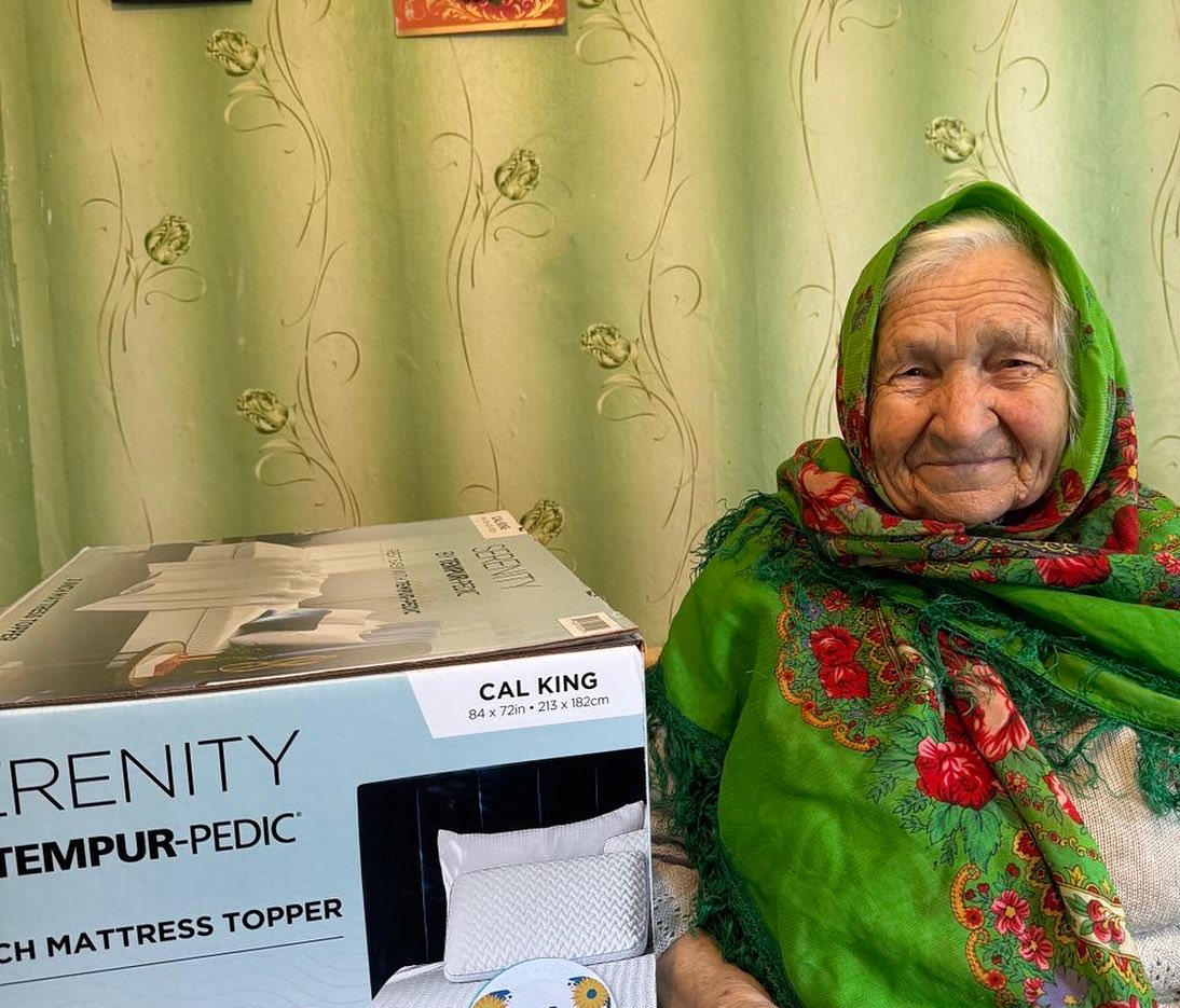 An elderly woman with a green headscarf smiling beside a boxed mattress topper.