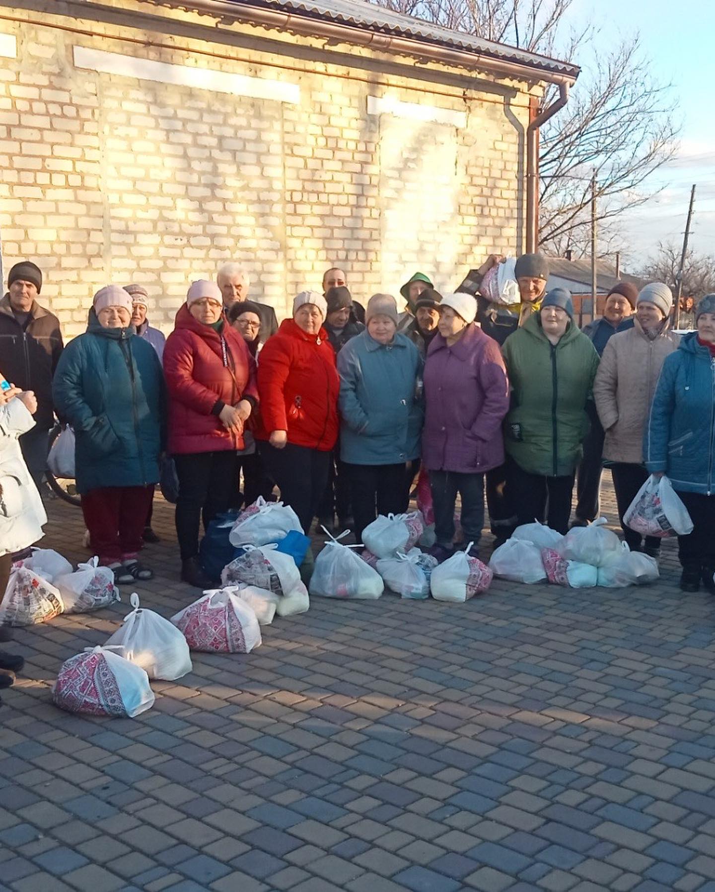 Group of elderly people standing outdoors in winter clothing with bags of groceries at their feet.