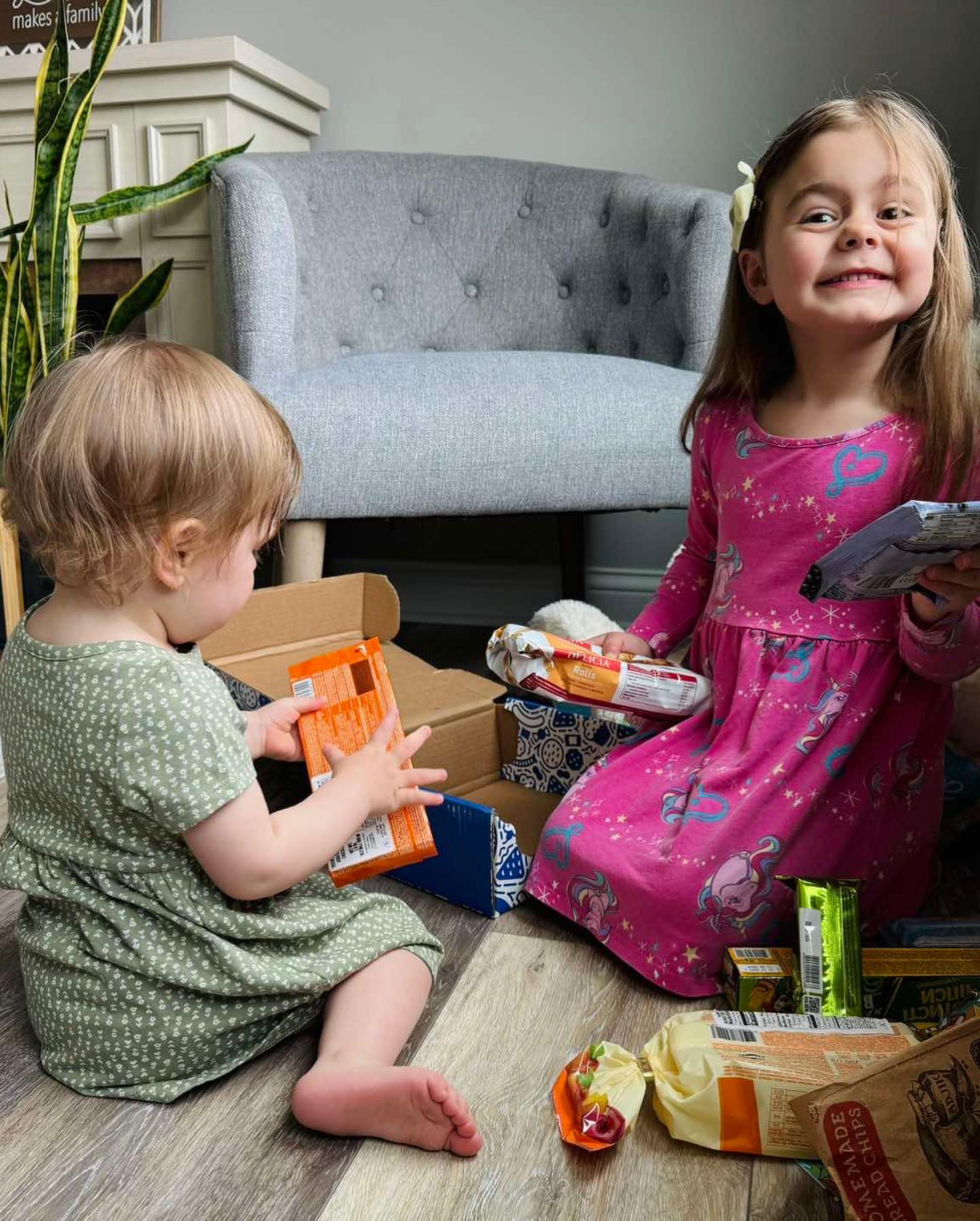 Two young girls sitting on the floor, playing with snack bars and packages, with a grey armchair and a plant in the background.
