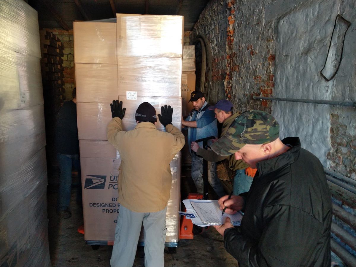 Men loading or unloading large cardboard boxes from a truck, with one man checking a clipboard.