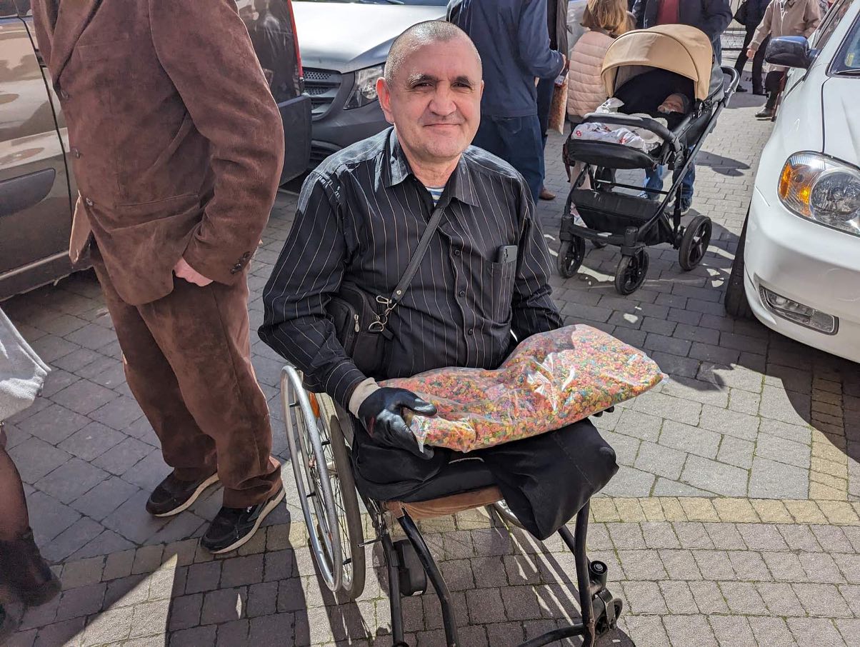 An elderly man in a wheelchair smiles at the camera, holding a colorful gift on his lap, surrounded by people outdoors.