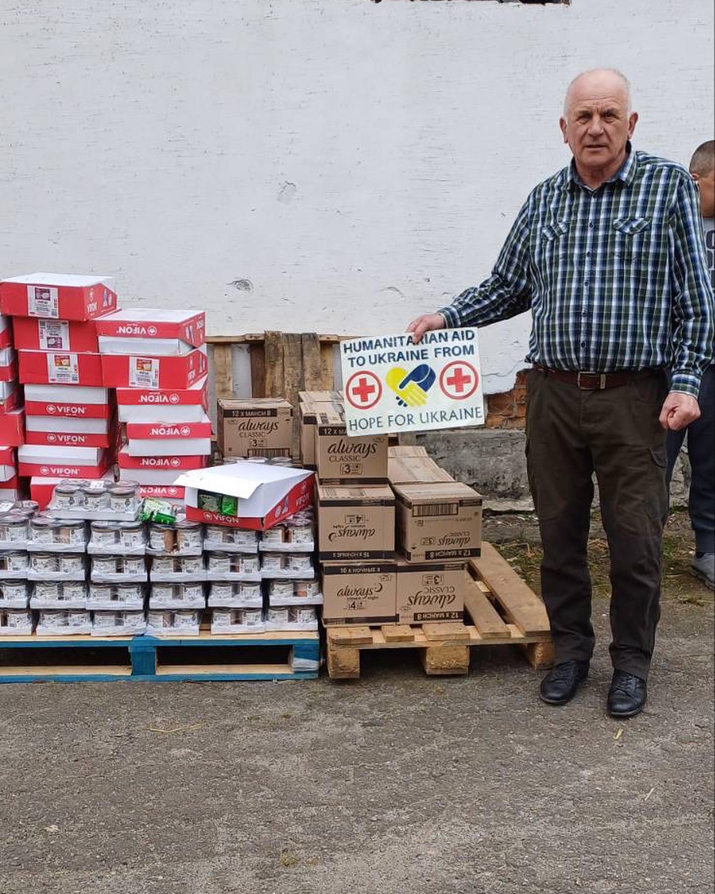 An elderly man standing beside boxes labeled "humanitarian aid to ukraine from hope for ukraine" and other supplies.